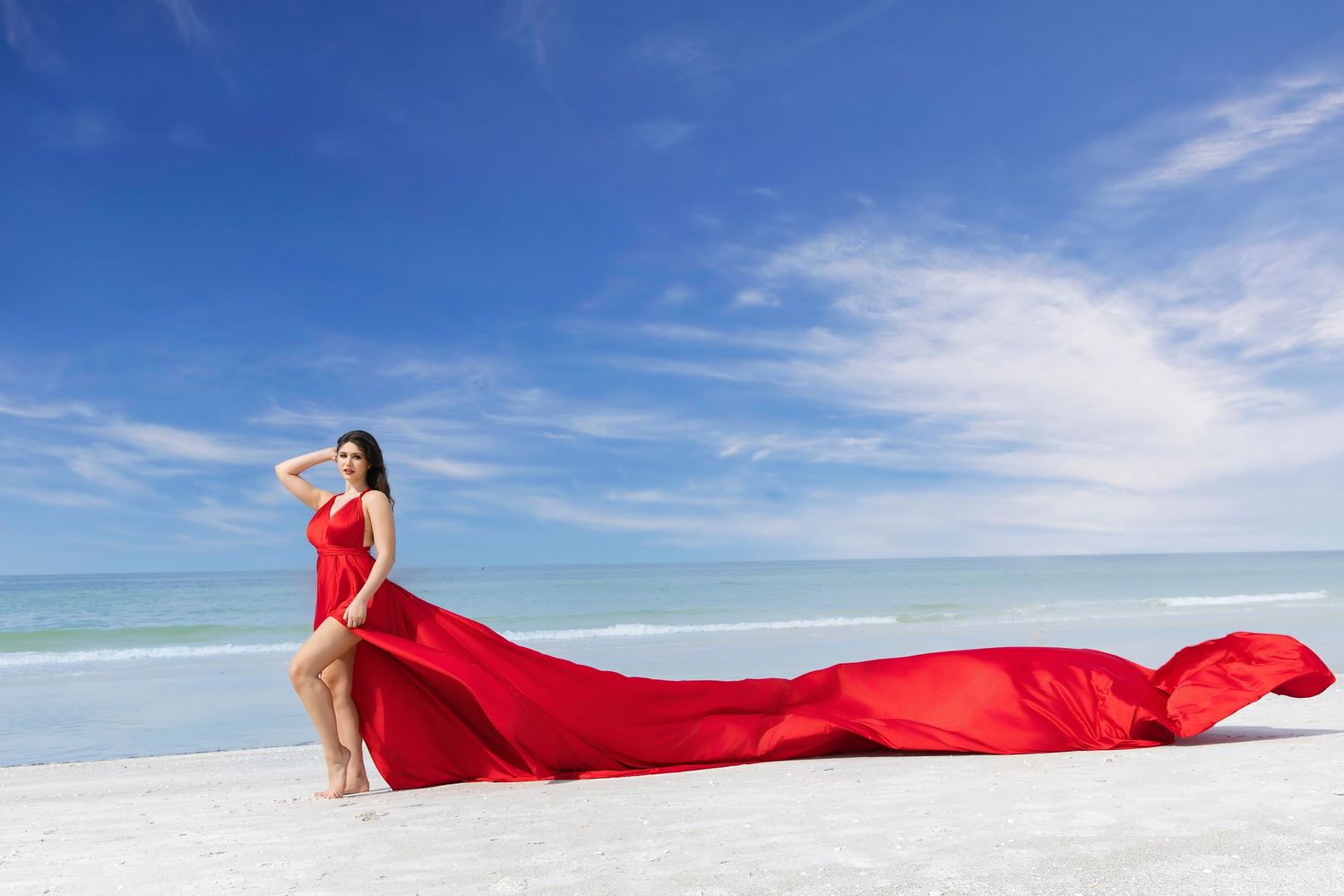 A flowing red fabric billows gracefully against a pristine white sand beach and bright blue ocean backdrop. Flying Dress Photoshoot