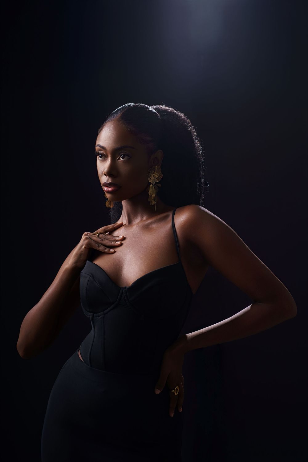 Dramatic portrait against dark background shows elegant figure in black dress with gold earrings striking a posed stance.