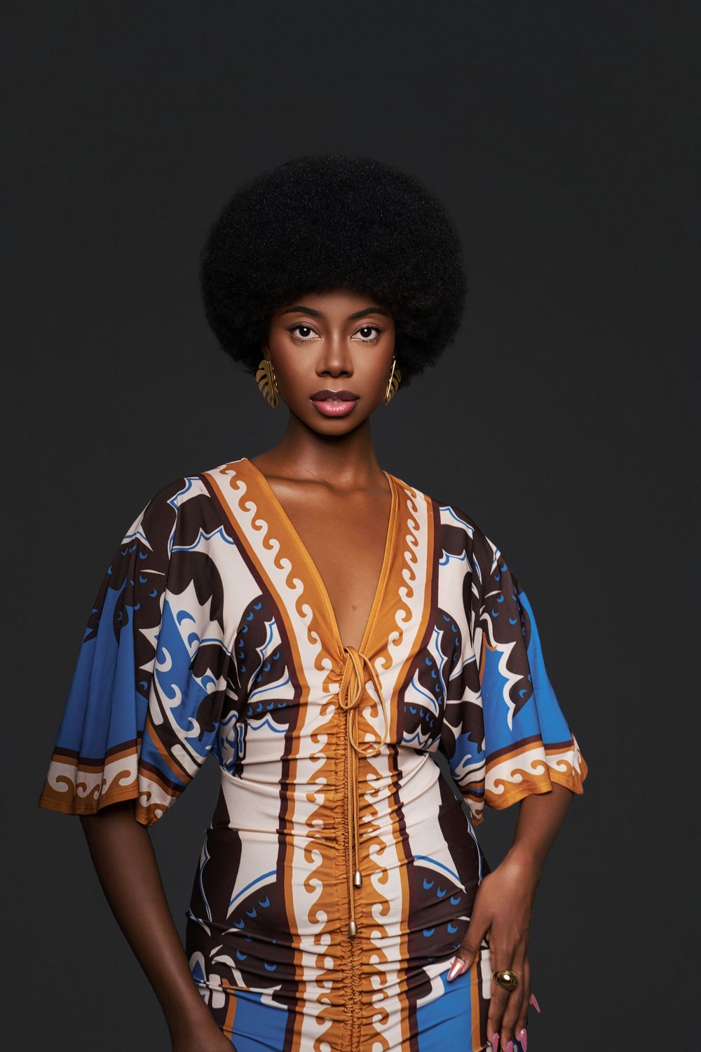 Fashion portrait of model wearing colorful patterned dress with orange accents and natural afro hairstyle against dark background.