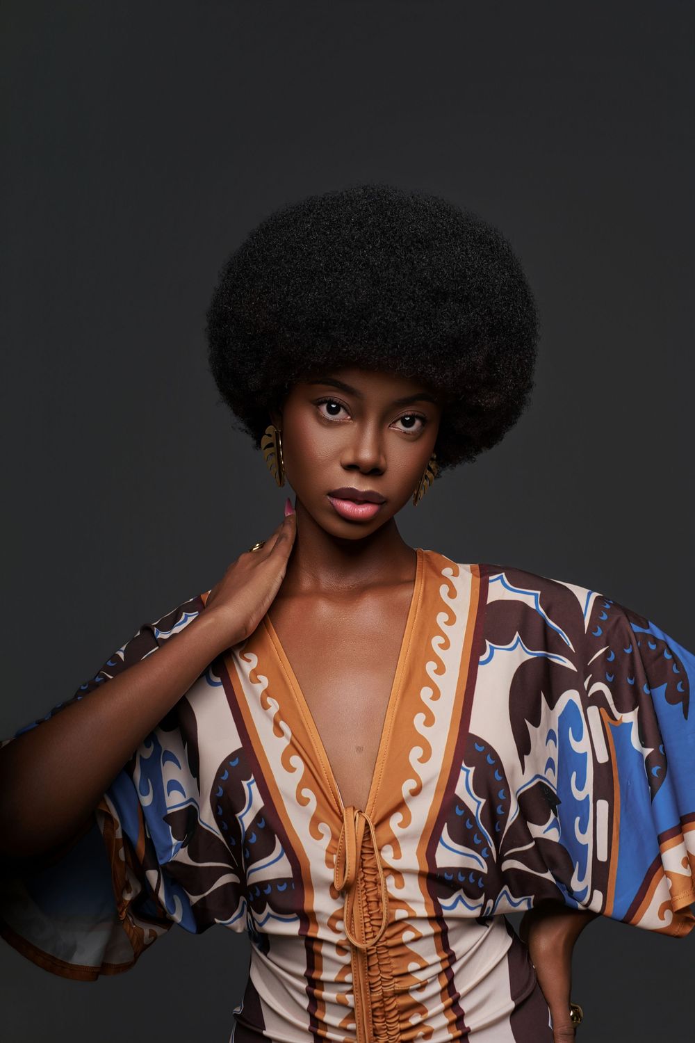 Fashion portrait of a model wearing a patterned dress with brown and blue geometric designs against a dark background.