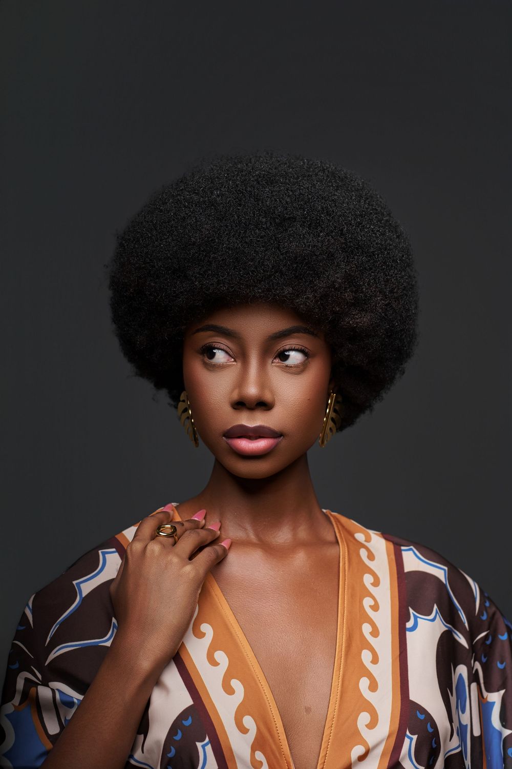 A person with natural curly hair wearing a patterned dress poses expressively against a dark background.
