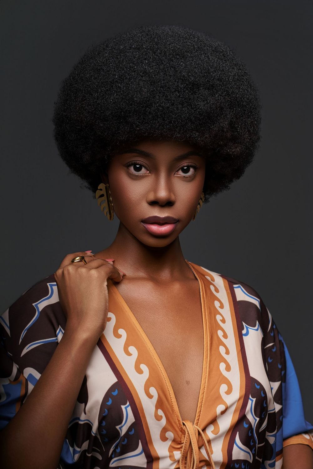 Stylish portrait featuring a natural afro hairstyle and patterned retro-style top with geometric designs against dark background.