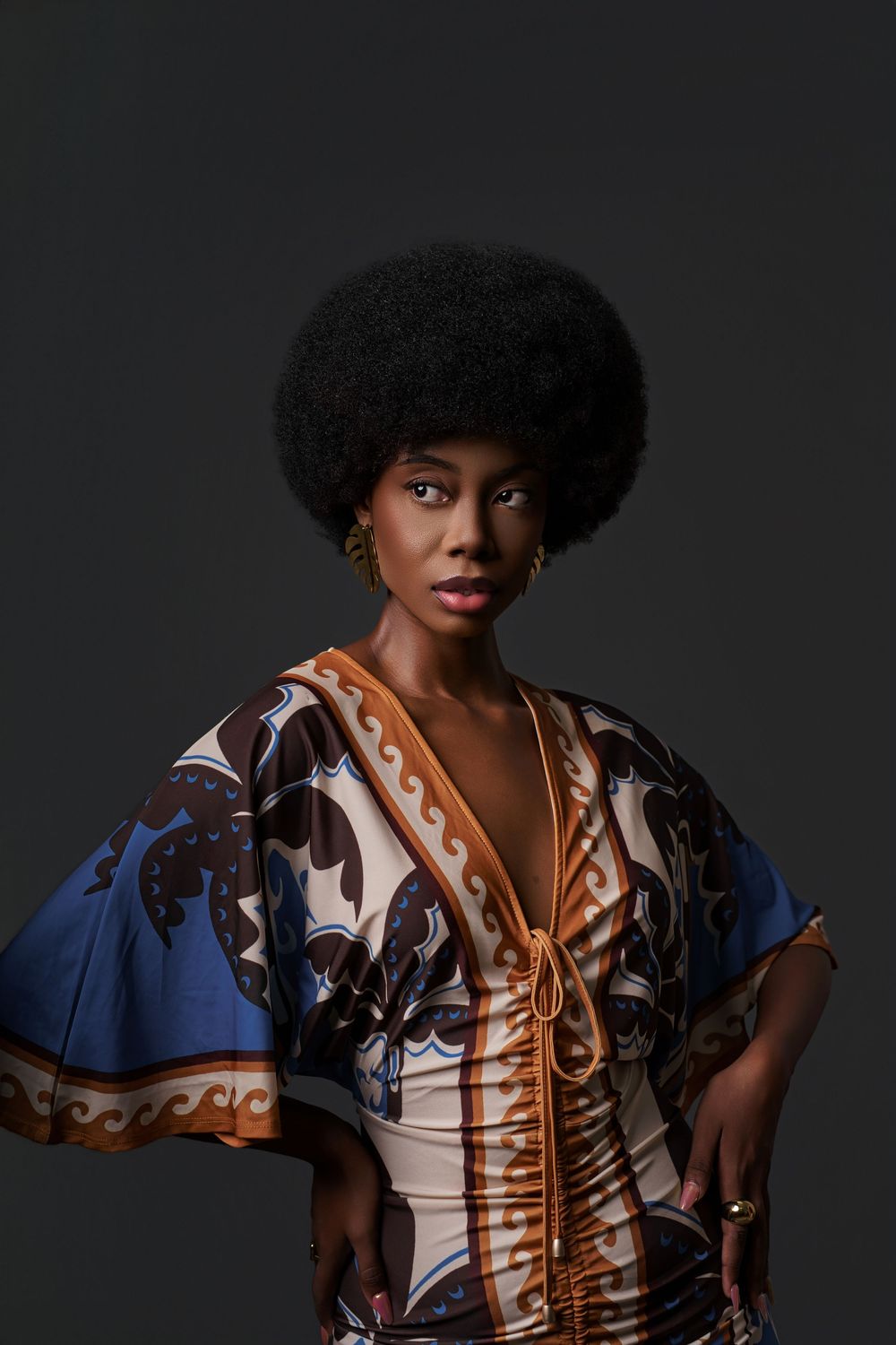 Fashion portrait of a model with natural hair wearing a patterned blue and brown wrap dress against dark background.