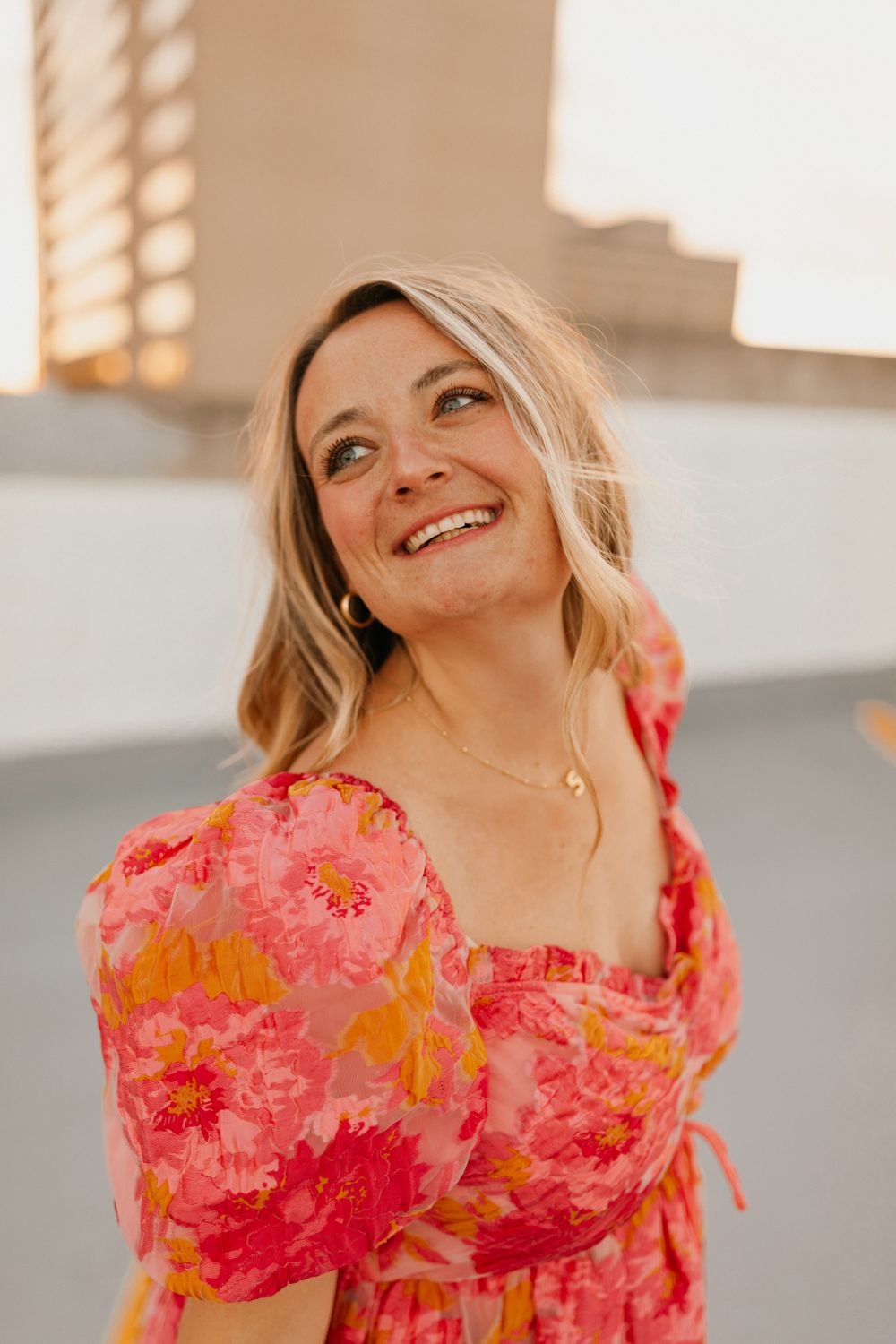 A girl in a coral pink floral dress smiles while posing against a light colored wall during golden hour lighting on a rooftop in Lexington.