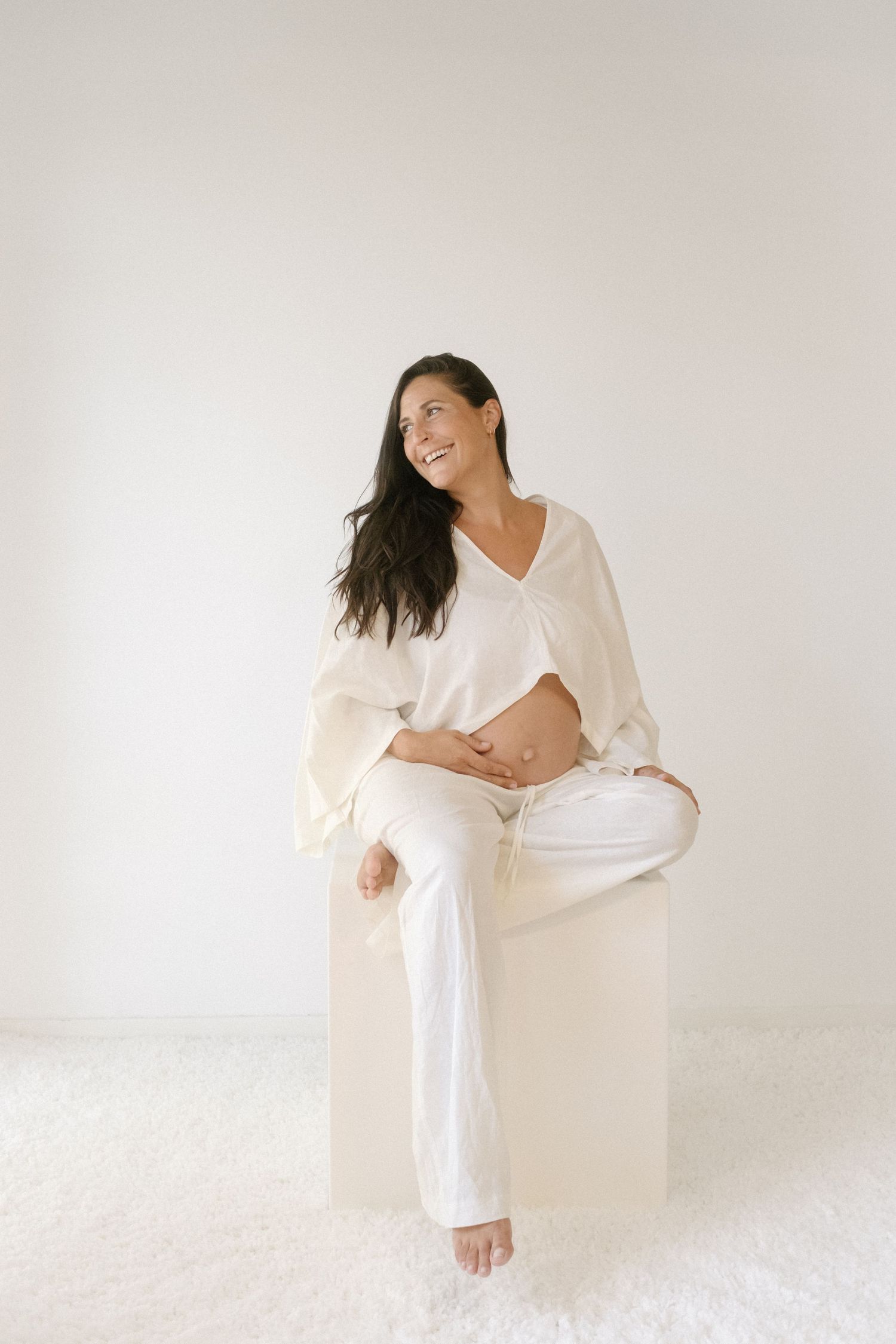 An expectant woman in white clothing sits on a white floor against a minimalist backdrop.