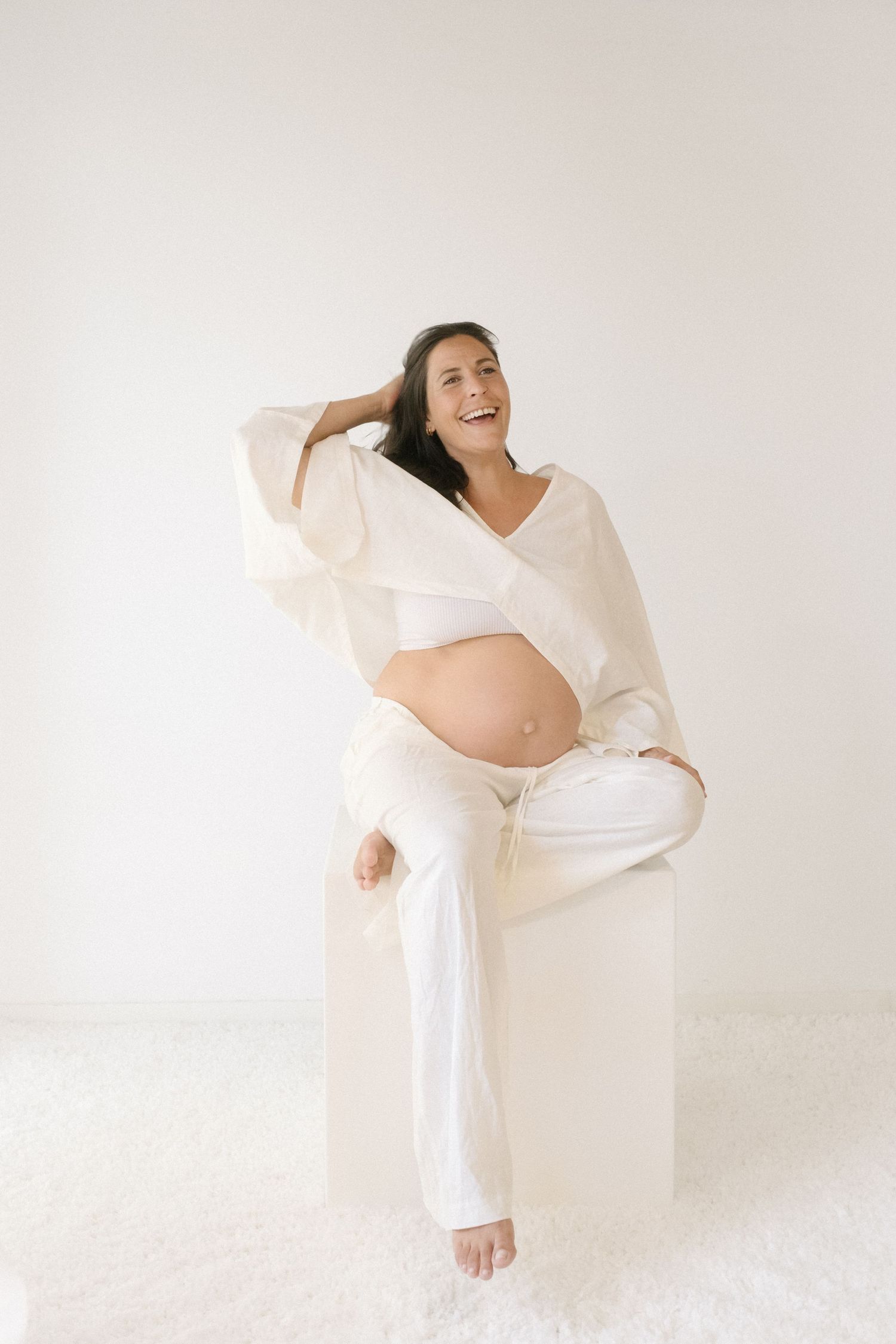 An expectant mother in white loungewear sits joyfully on a white pedestal against a minimalist background.