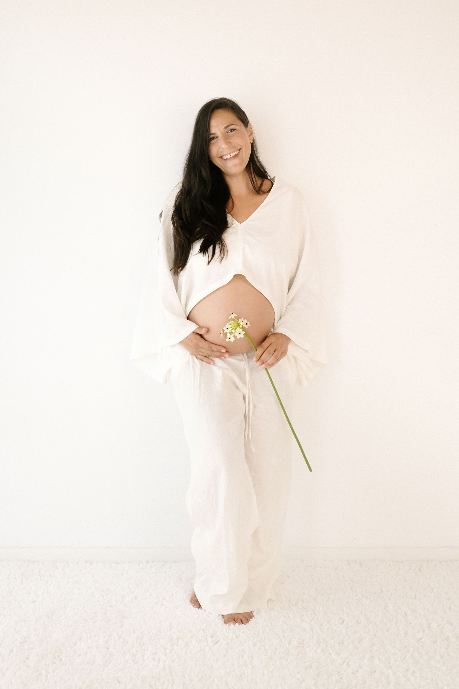 Expectant woman in white flowing outfit holds small flower against light background in minimalist maternity photo.
