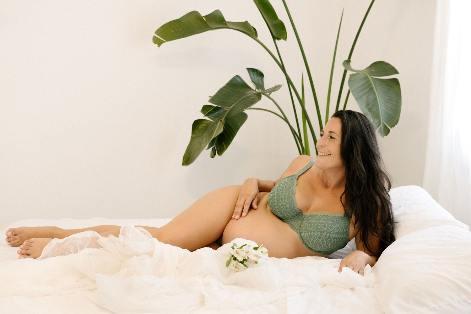 Woman in green lingerie lounges on white bedding with tropical plants in the background.