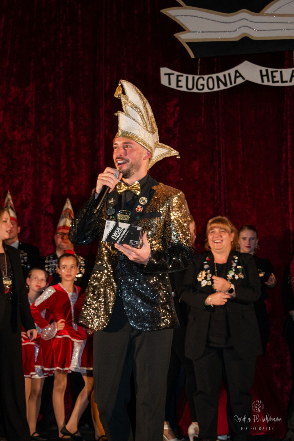 A performer in a gold and black costume holds a ceremonial horn on stage with choir members in the background.