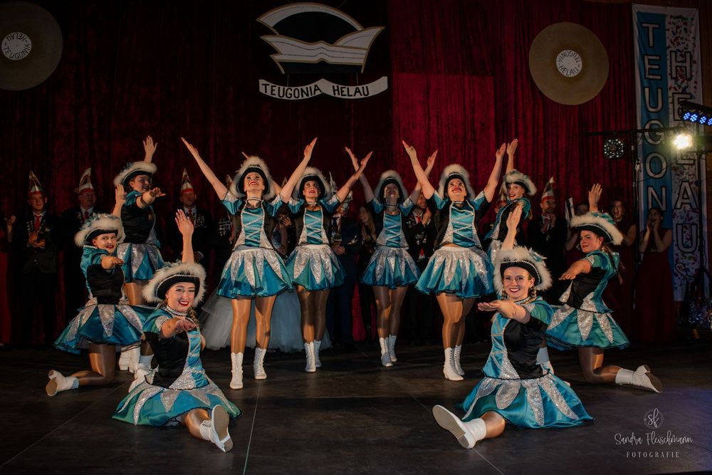 Ballet dancers in turquoise tutus perform a synchronized split leap sequence on a dark stage during a recital.