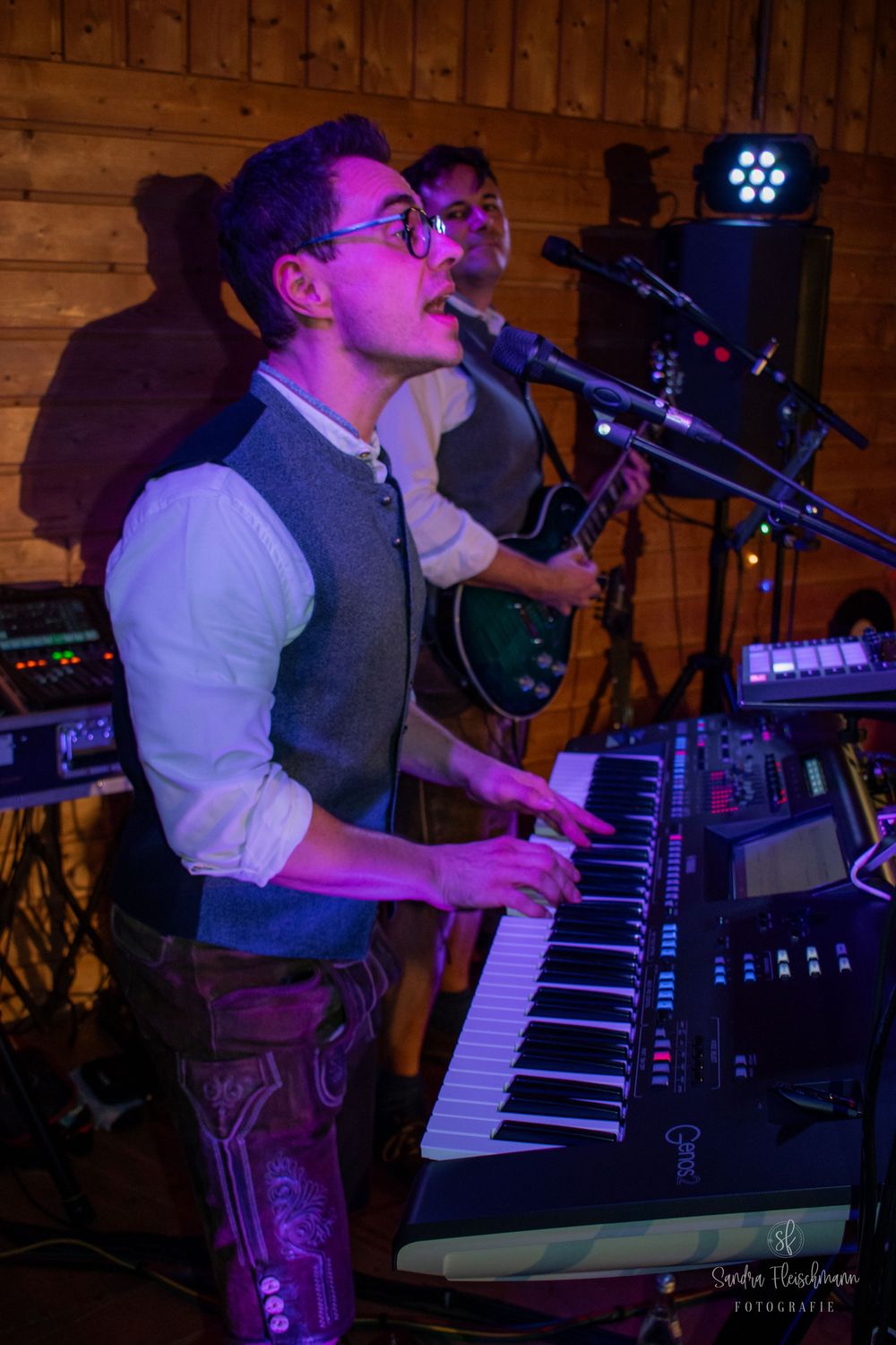 Musician in formal attire performs energetically on keyboard at dimly lit venue with purple stage lighting.