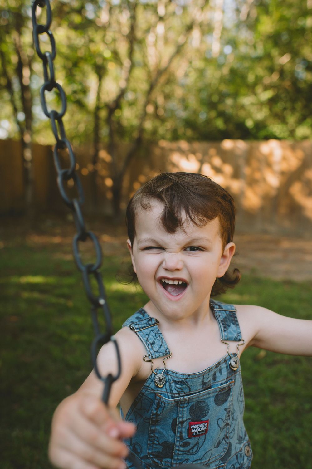 A joyful child in blue overalls plays on a swing in a sunny backyard.