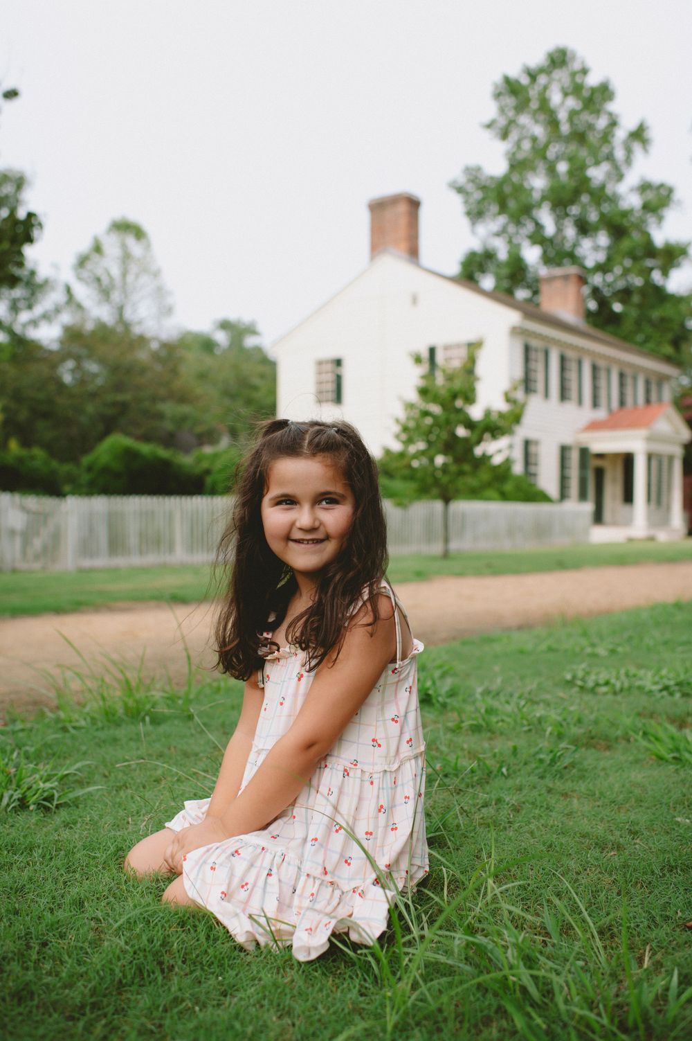 A summer portrait of a young person in a white dress sitting on grass near a historic white house.