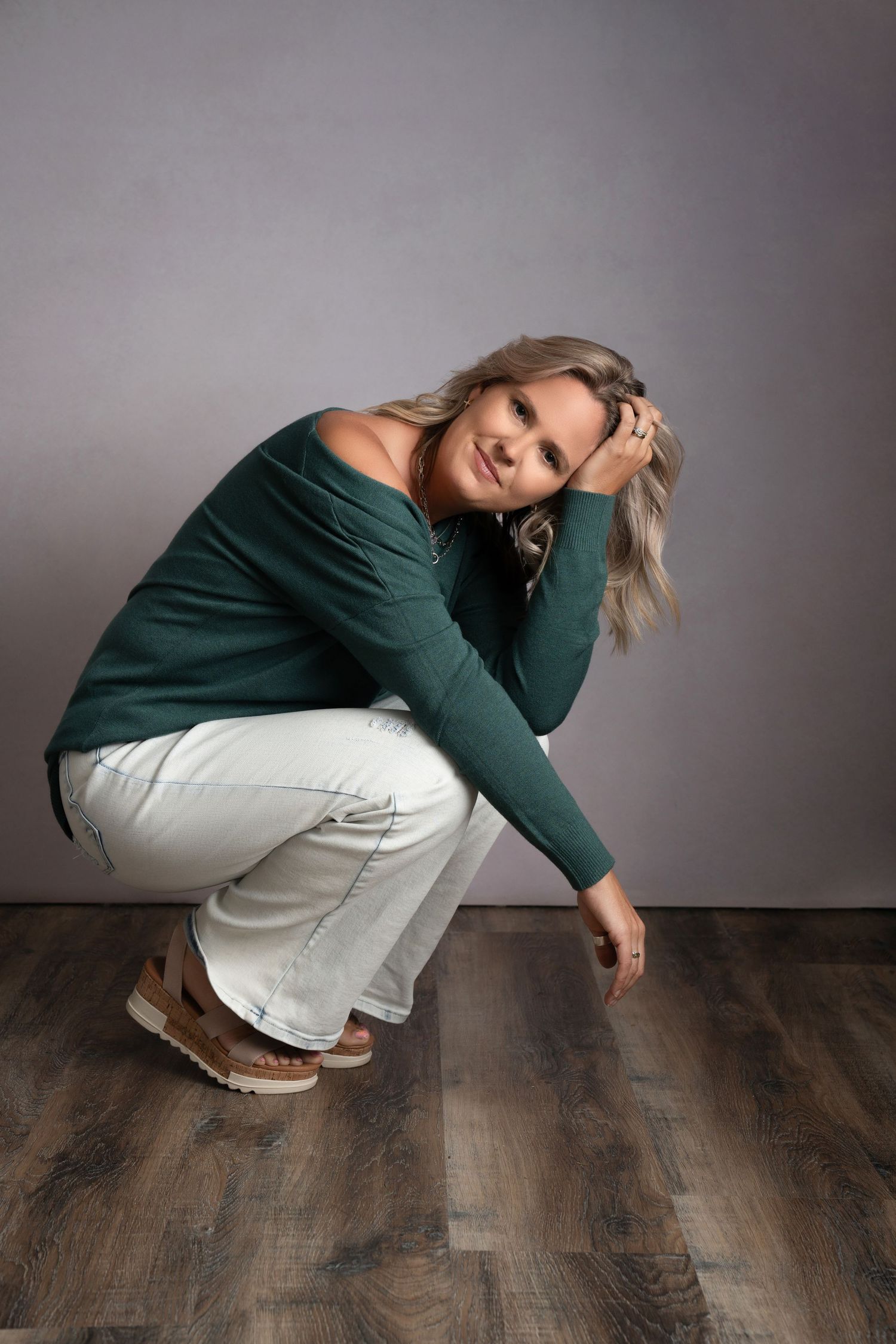 Woman in emerald green top and white pants poses against white wall in minimalist fashion series.