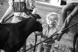 A young person in western attire interacts with a black horse at an equestrian event in black and white photography.