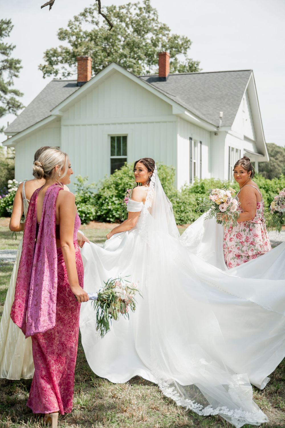 Wedding party helping with bride's dress outside white farmhouse buildings.
