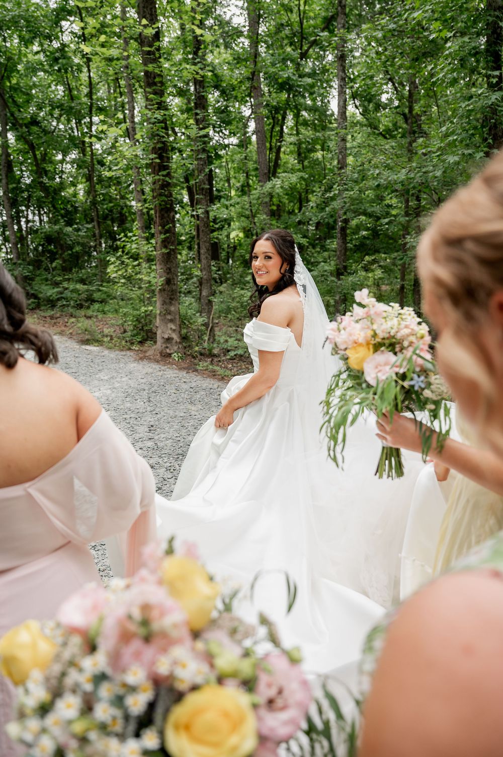 Wedding party gathered around bride's flowing dress in wooded setting.