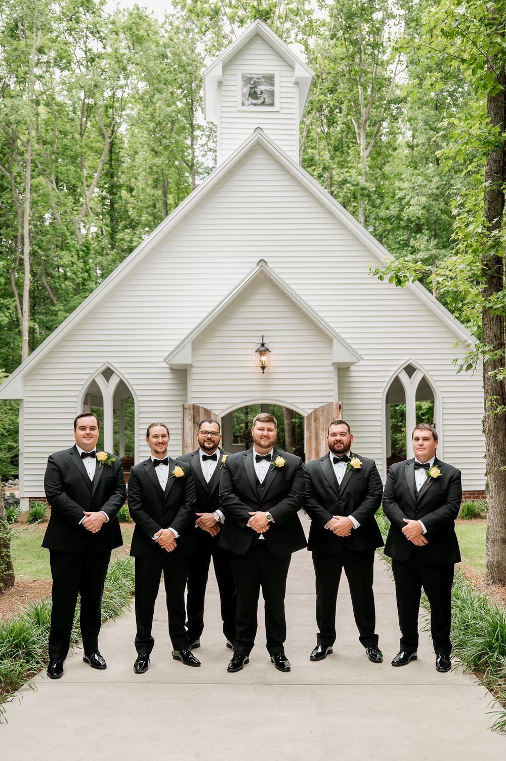 Groomsmen in black tuxedos pose in a line outside a white chapel on a wedding day.