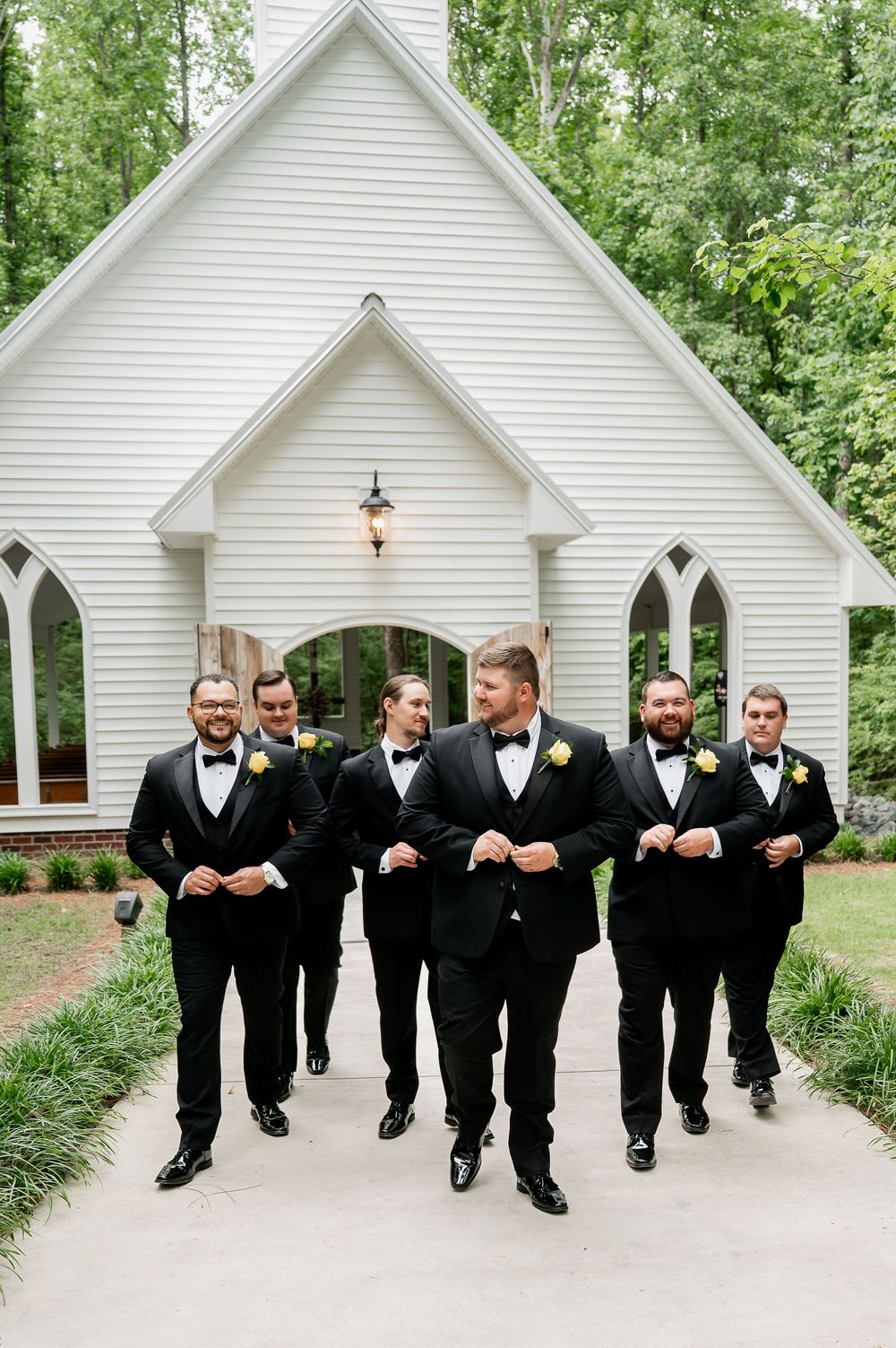 Wedding party members in black suits walk together on path in front of white church.