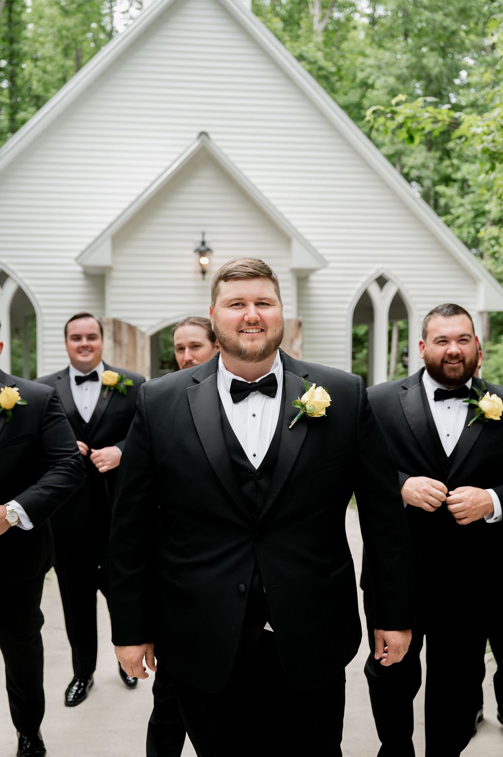 Groomsmen in formal black tuxedos gather for candid photo outside white chapel.