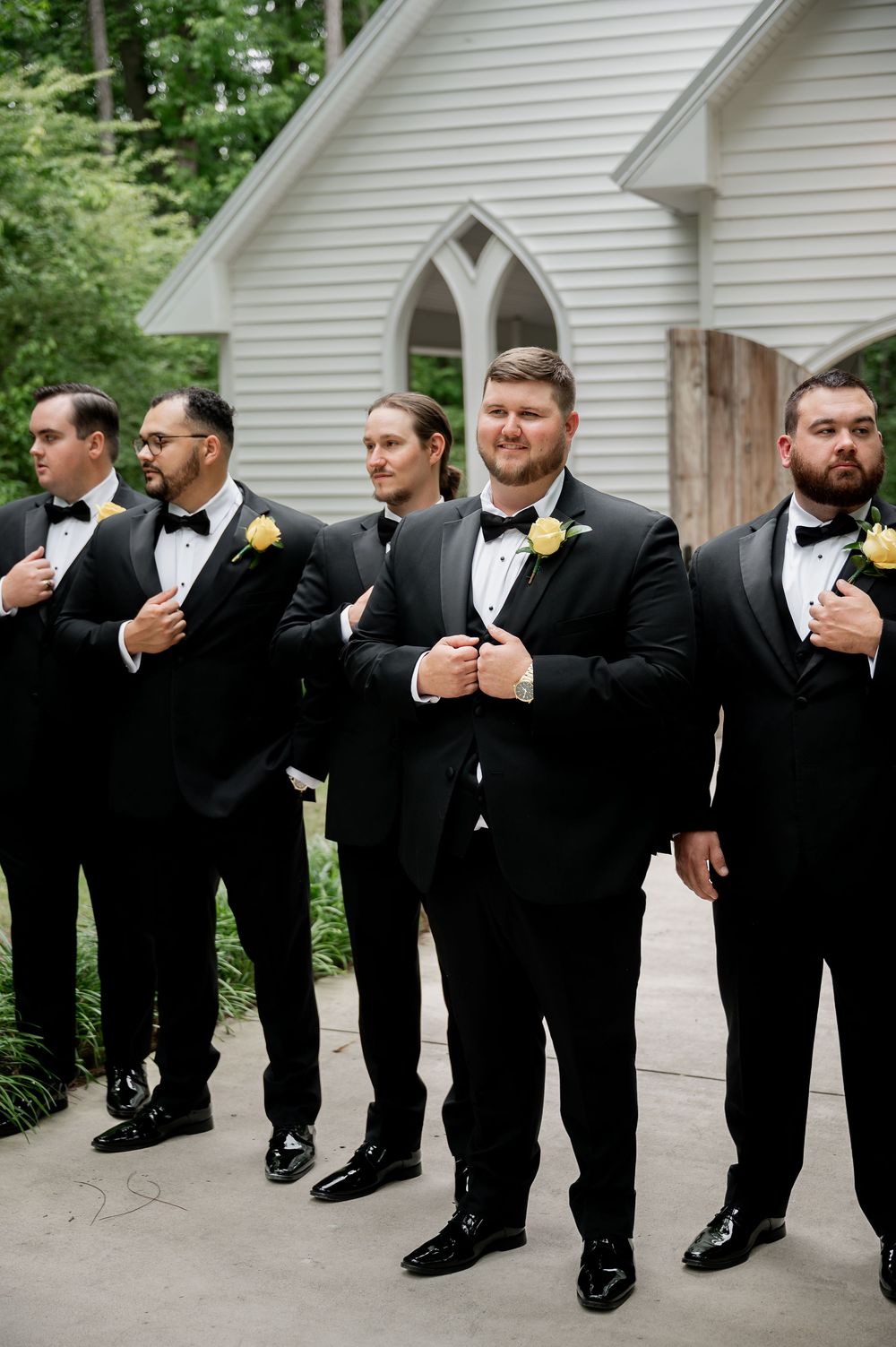 Wedding party members in black suits pose casually in front of church entrance.