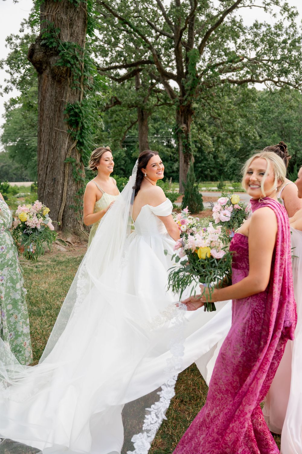 Wedding party helping with the bride's flowing white dress train in a garden setting.