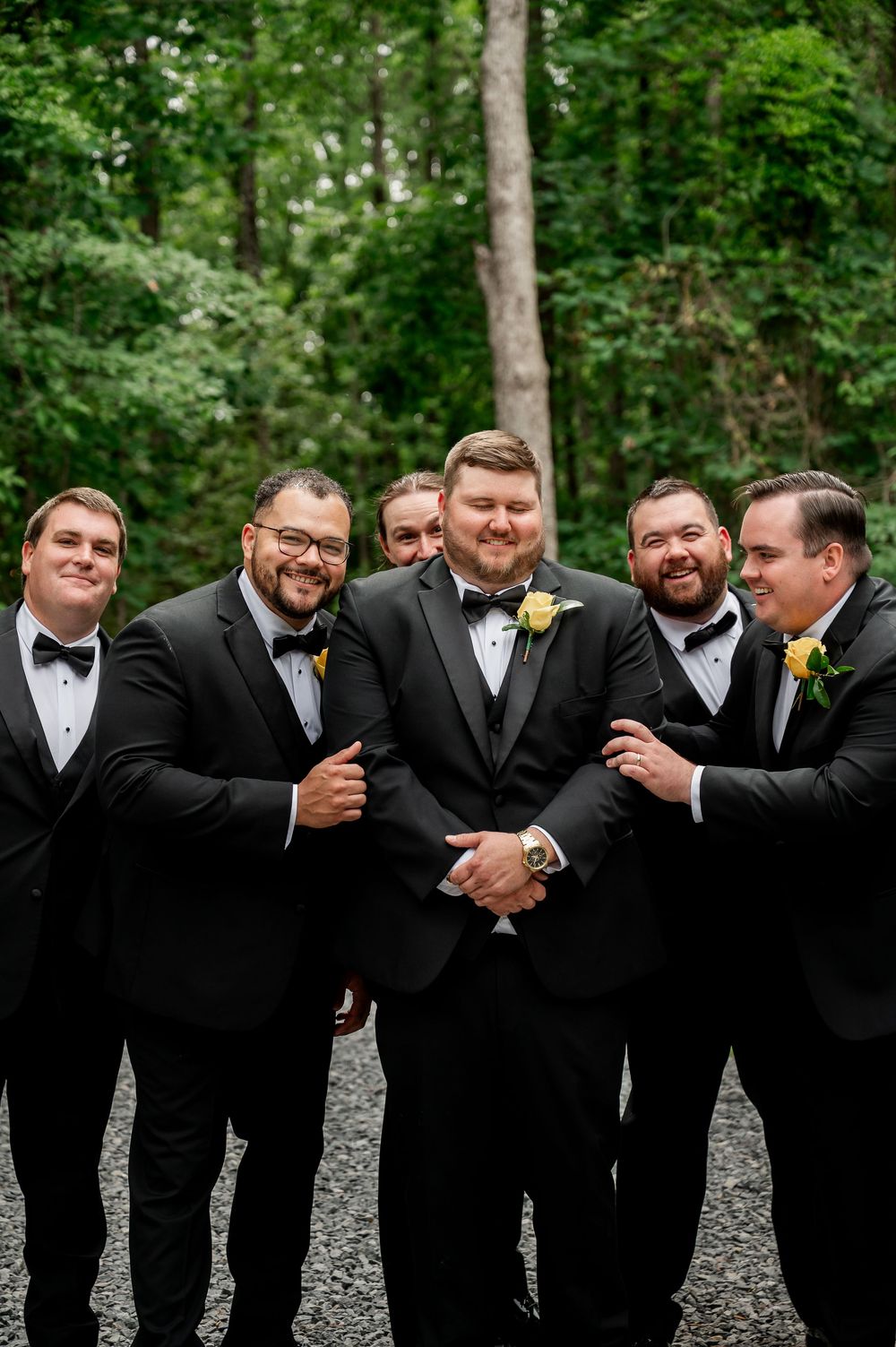 Groomsmen share laughs while posing for casual photos at outdoor wedding venue.
