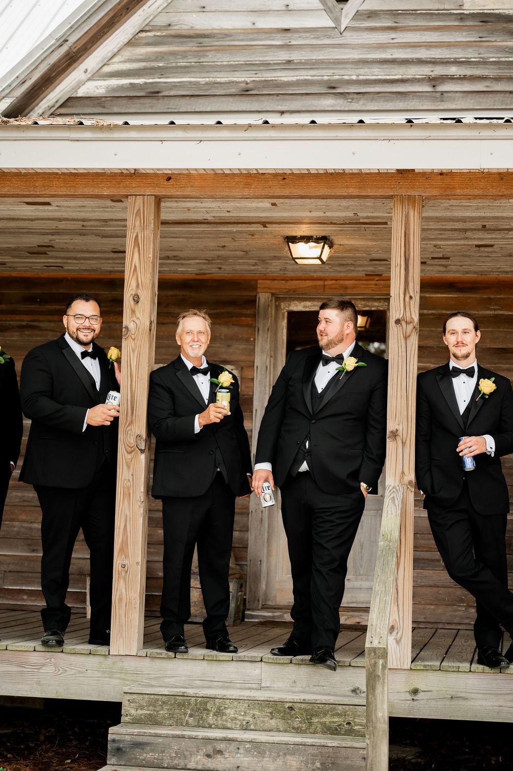 Groomsmen in black tuxedos pose on rustic wooden porch with drinks.