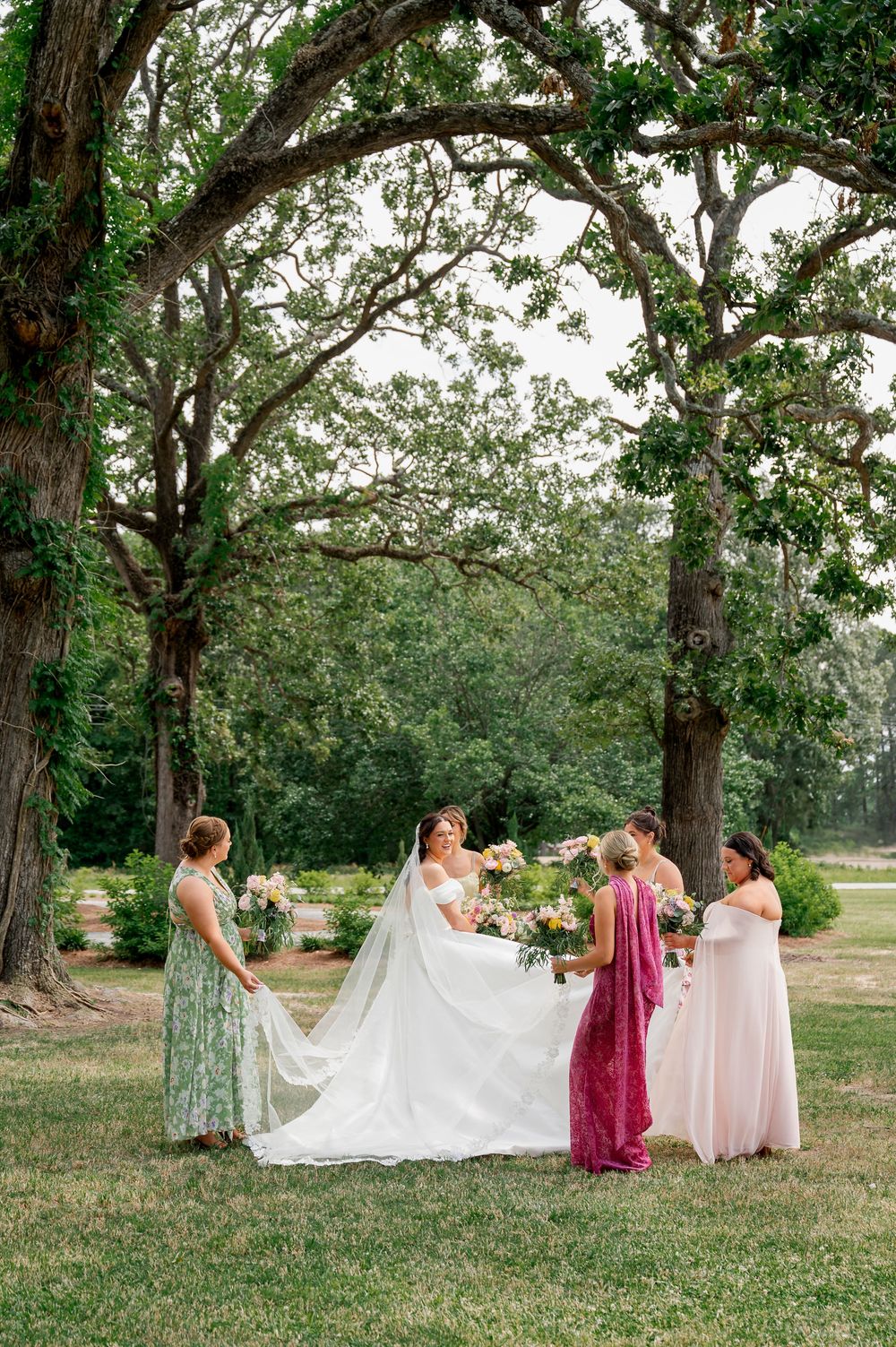 Wedding party arranging the bride's dress for photos under large oak trees.
