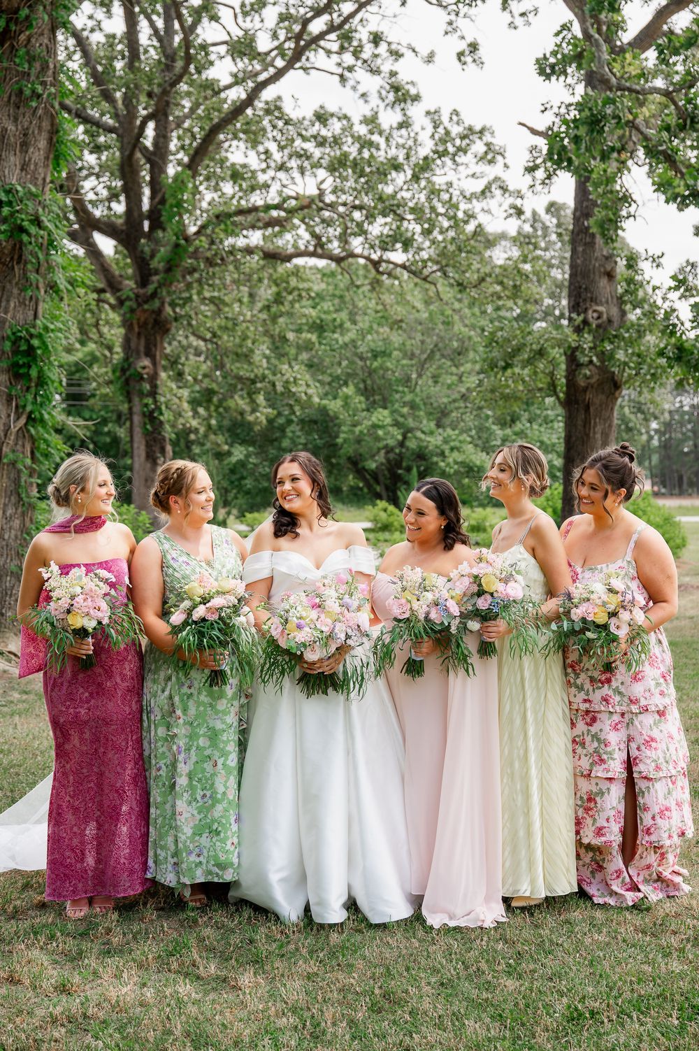Bridesmaids in floral and solid-colored dresses holding bouquets pose together outdoors.