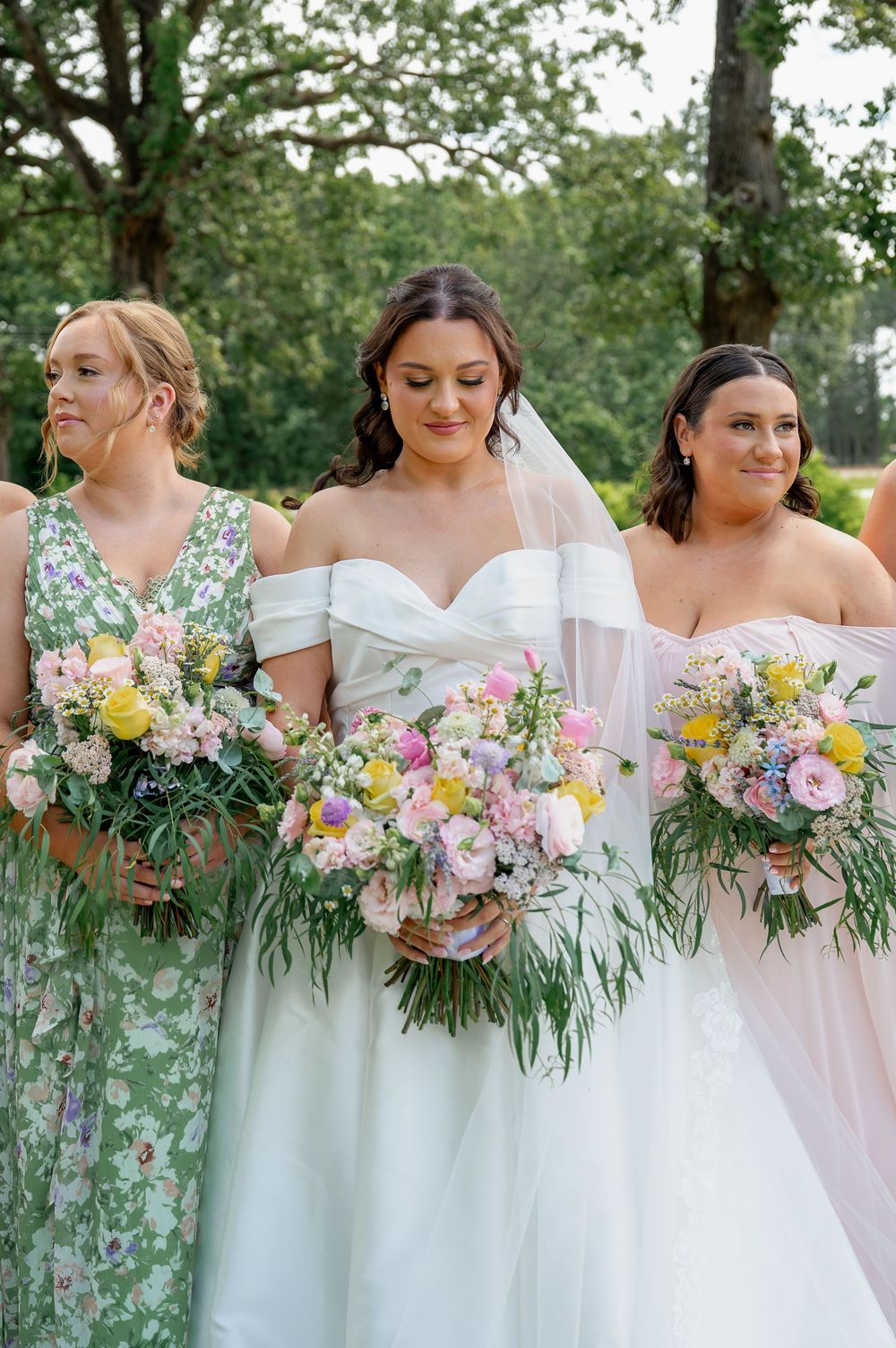 Row of bridesmaids holding pink and yellow floral bouquets against green foliage.