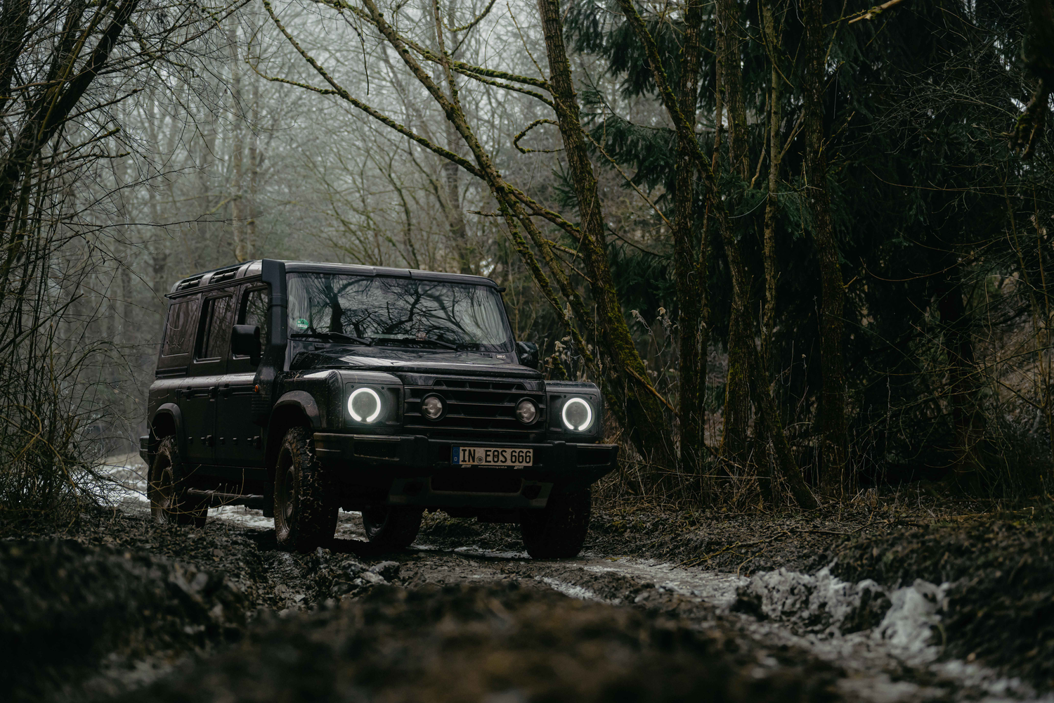 Black Ineos Grenadier driving through a misty forest path with patches of snow on the ground.