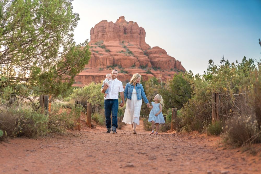 People walking along desert trail with towering red rock formations in background.