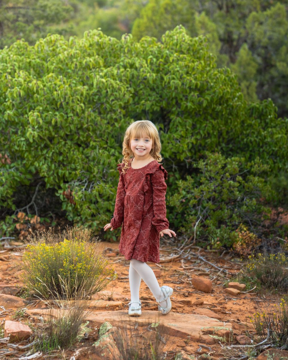 A young child in a burgundy dress twirls playfully against a backdrop of green shrubs and red desert soil.