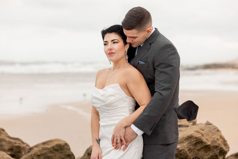 A couple in wedding attire embrace on rocky beach during overcast day, with waves in background.