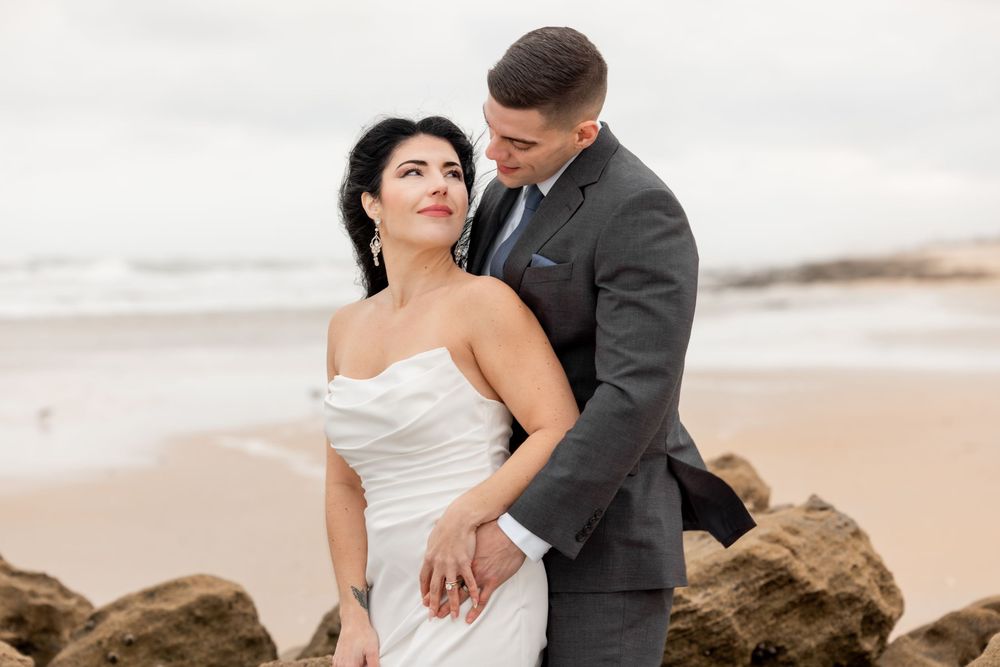 A romantic couple in formal wedding attire embrace on a sandy beach near rocky outcrops during sunset.
