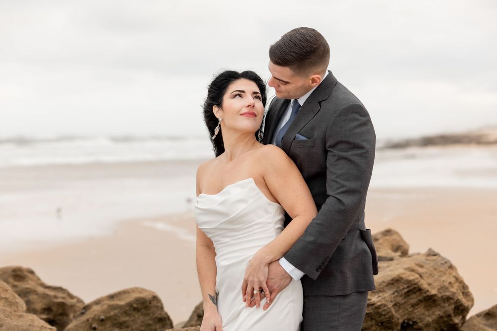 A romantic couple in wedding attire embraces on a scenic beach with waves and rocks in the background.