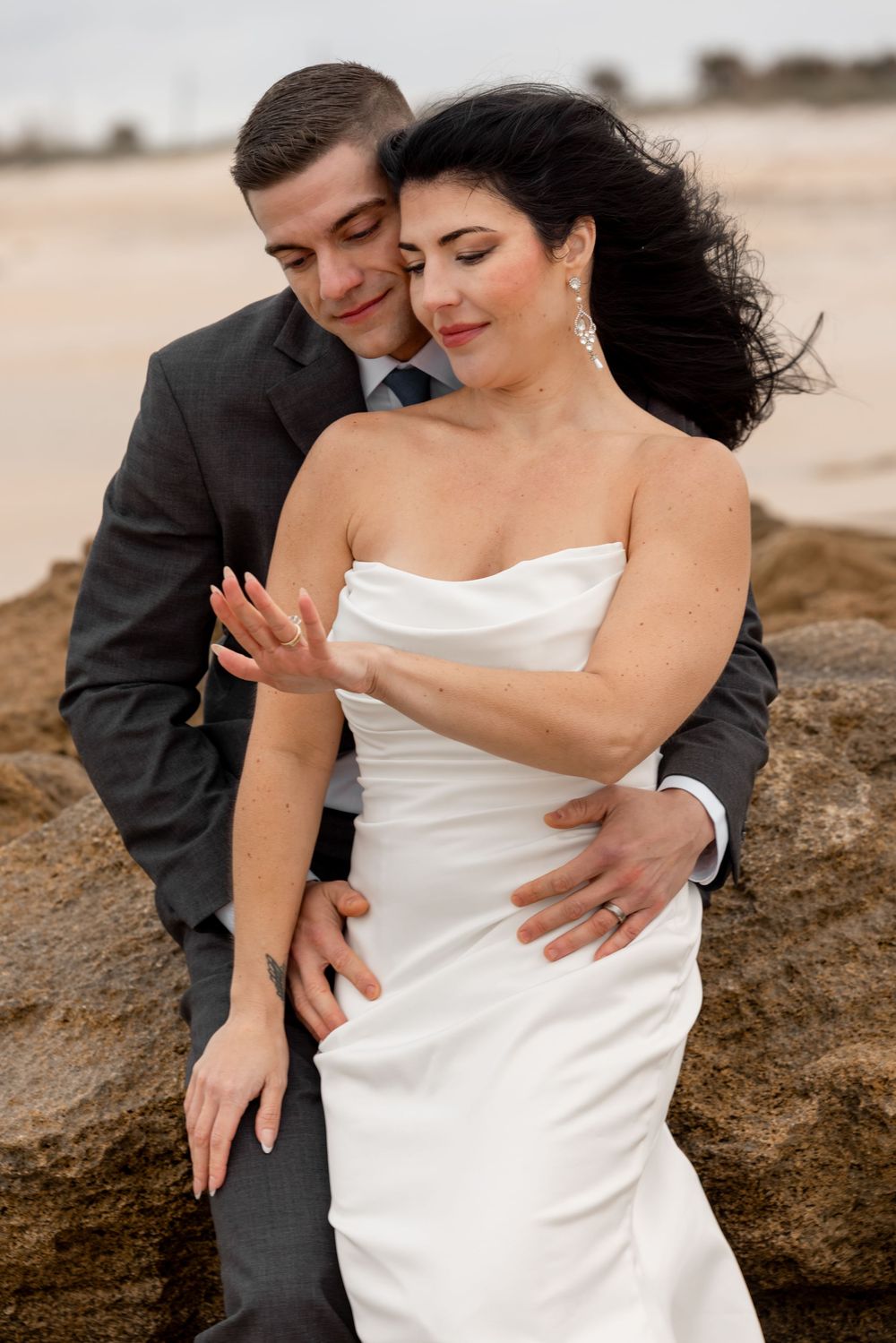 Romantic beach wedding photos showing couple embracing on rocky shore in elegant attire.