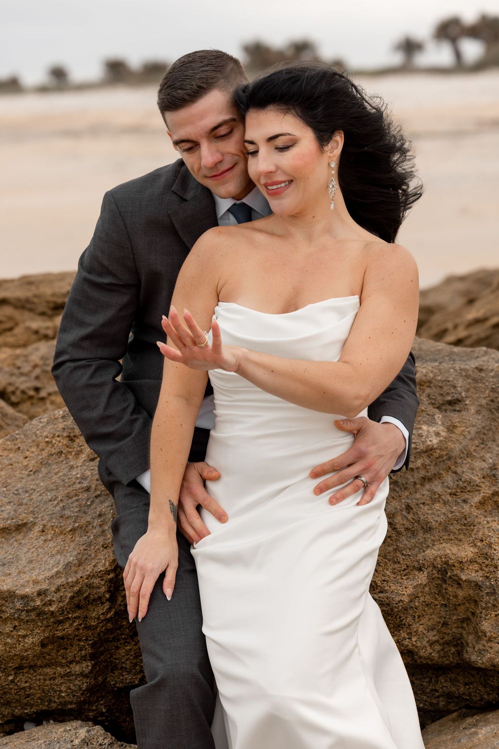 A couple embraces on rocky coastal terrain, with the bride wearing a white dress and groom in dark suit.