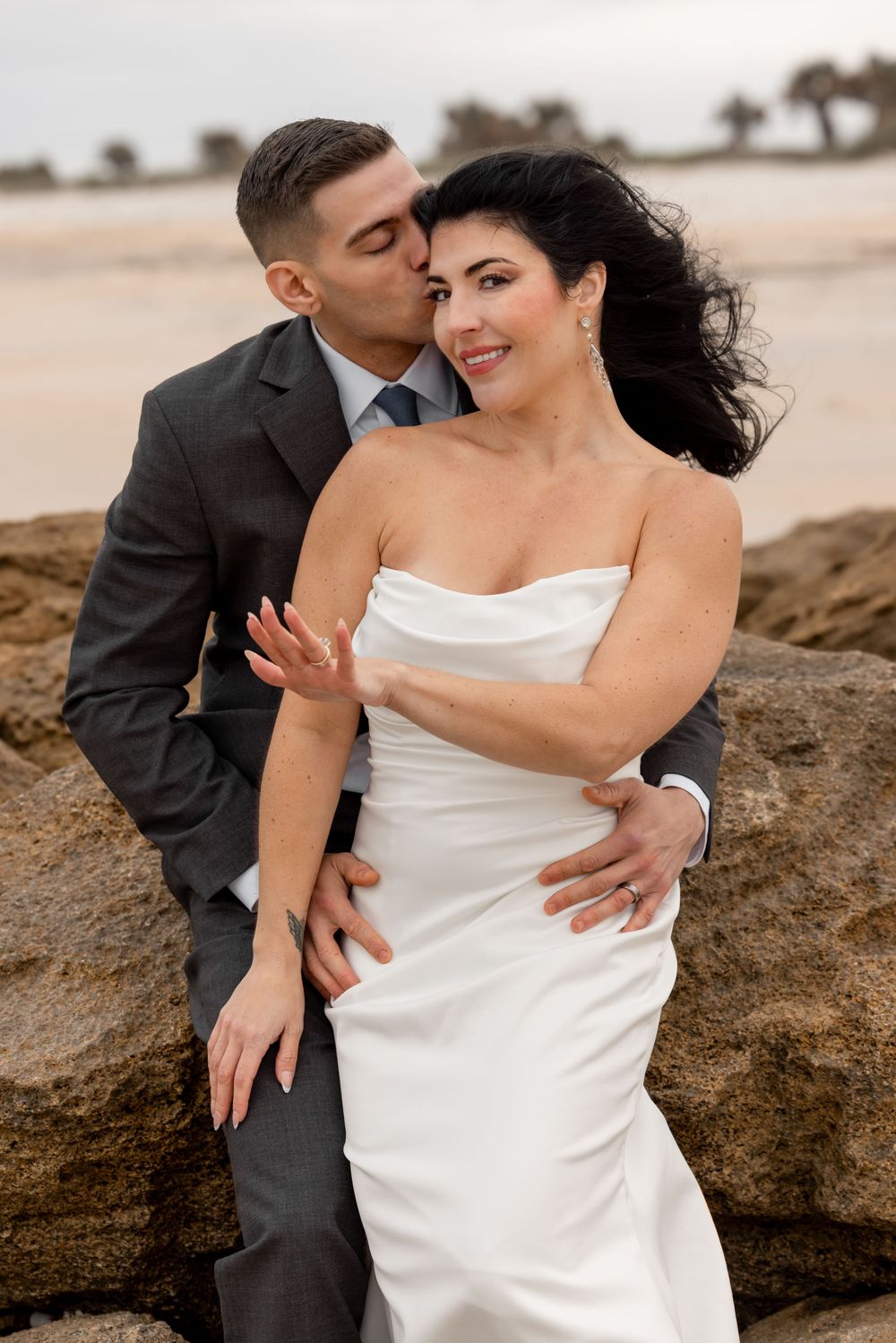 Romantic beach wedding portrait of a couple embracing on coastal rocks at sunset.