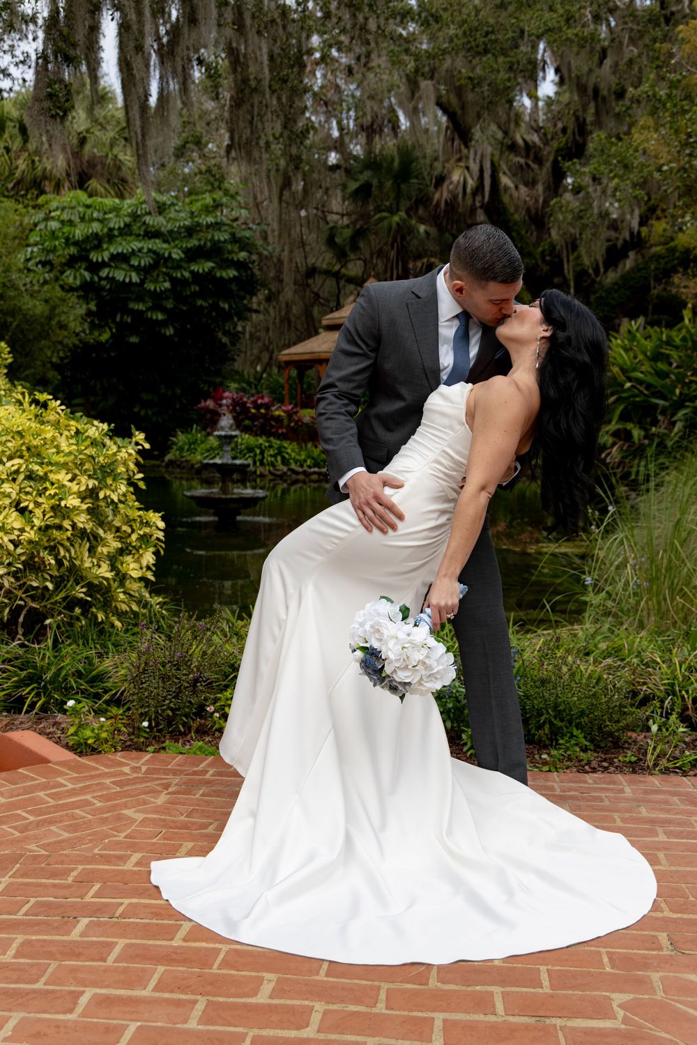 A romantic dip pose between a couple in formal wedding attire on a brick patio surrounded by lush gardens.