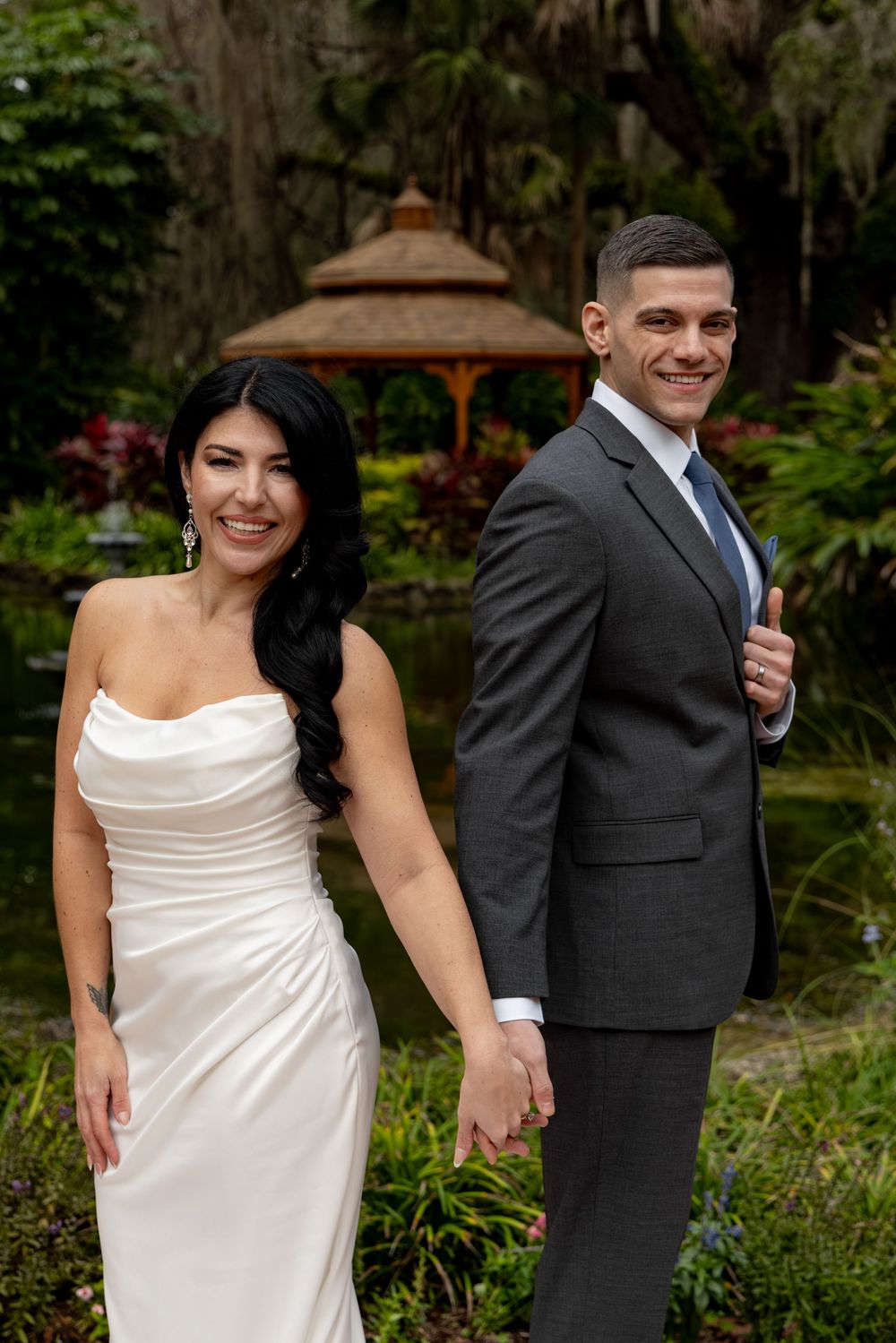 Wedding couple poses playfully in lush garden setting with gazebo in background.
