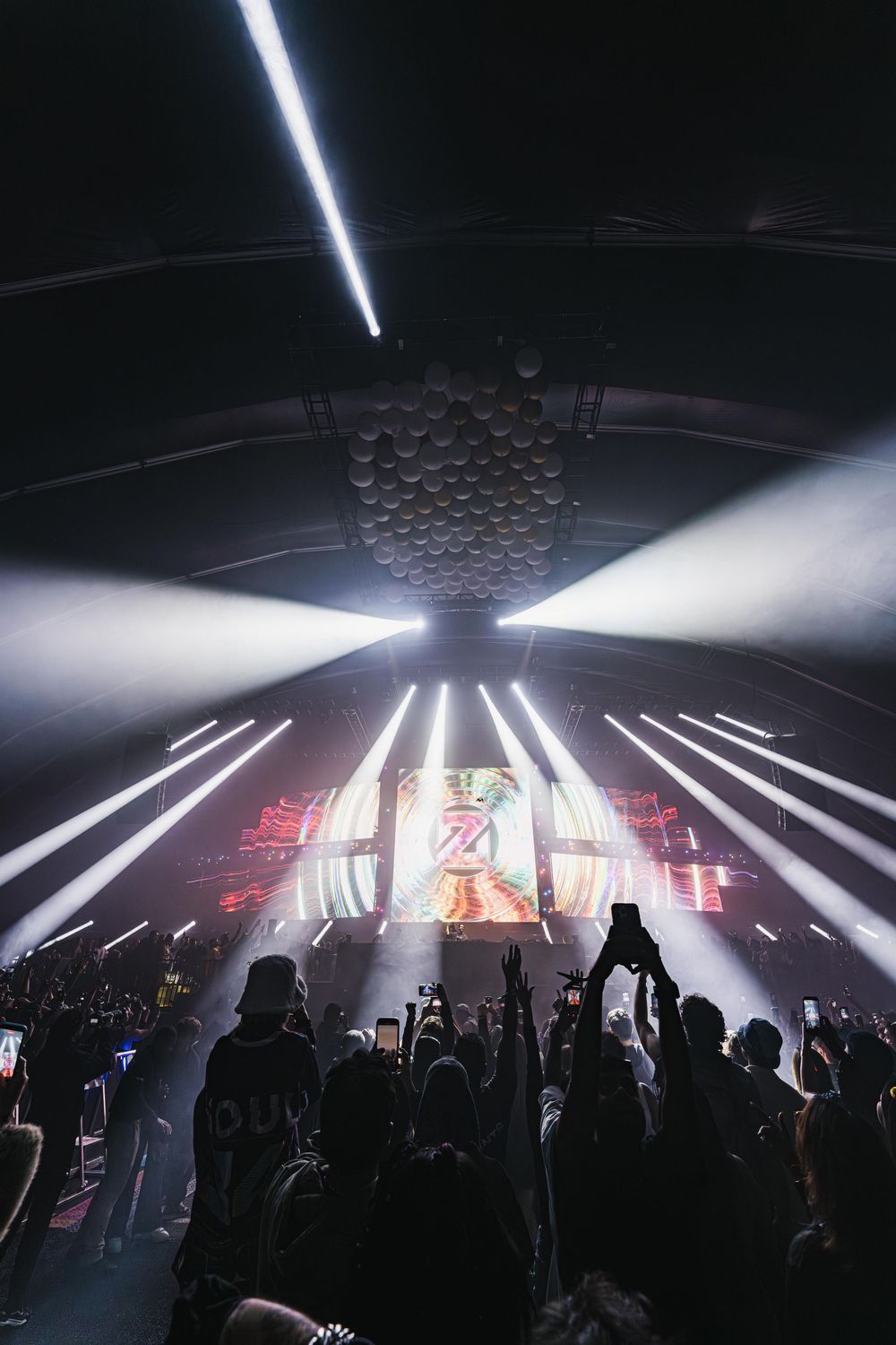 Concert crowd silhouettes in a dark venue with dramatic white beam lights creating geometric patterns overhead.
