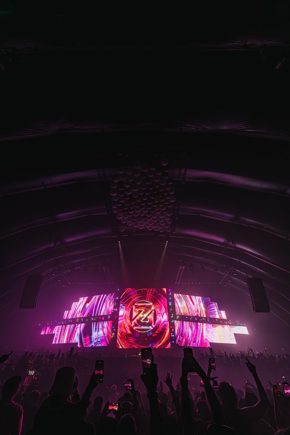 Concert crowd silhouettes against vibrant purple stage lighting and large LED display screens at an electronic music event.