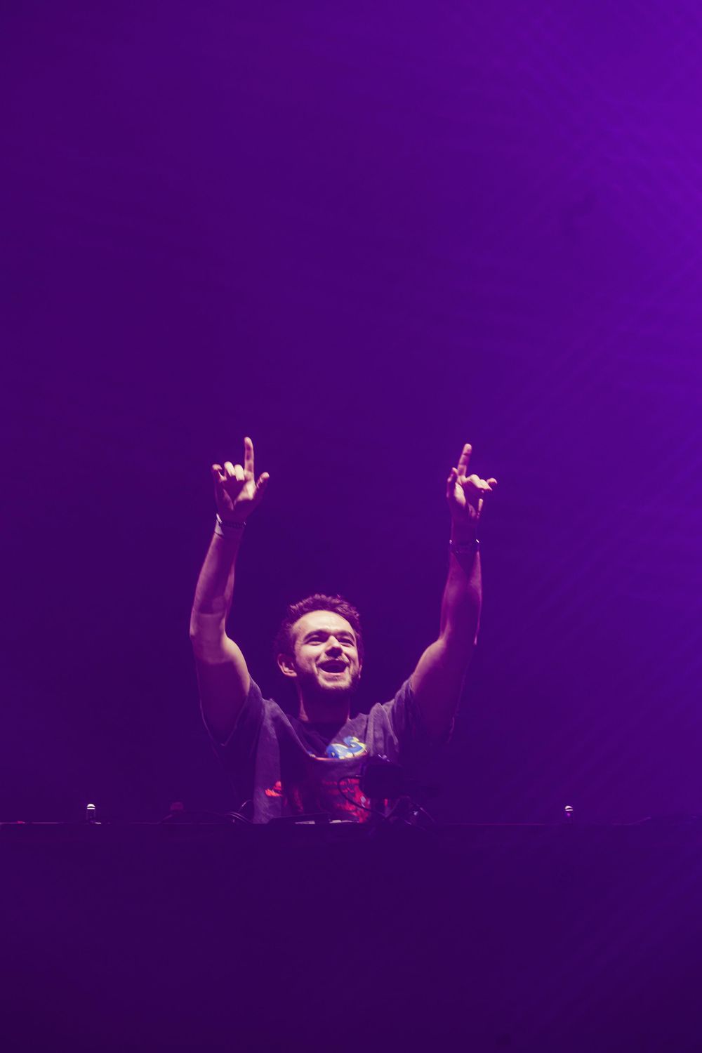 DJ performs behind decks on stage with raised arms in purple lighting at electronic music concert.