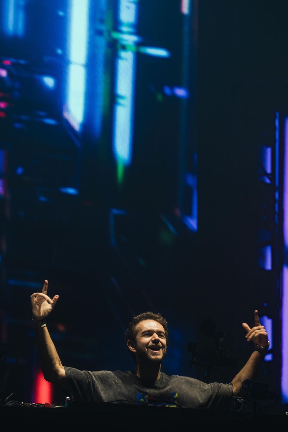 DJ performing on stage with raised arms against dark background with blue lighting effects and vertical light beams.