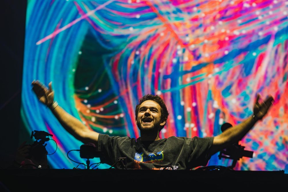 DJ performing at electronic music festival with vibrant blue and pink light show display in background.