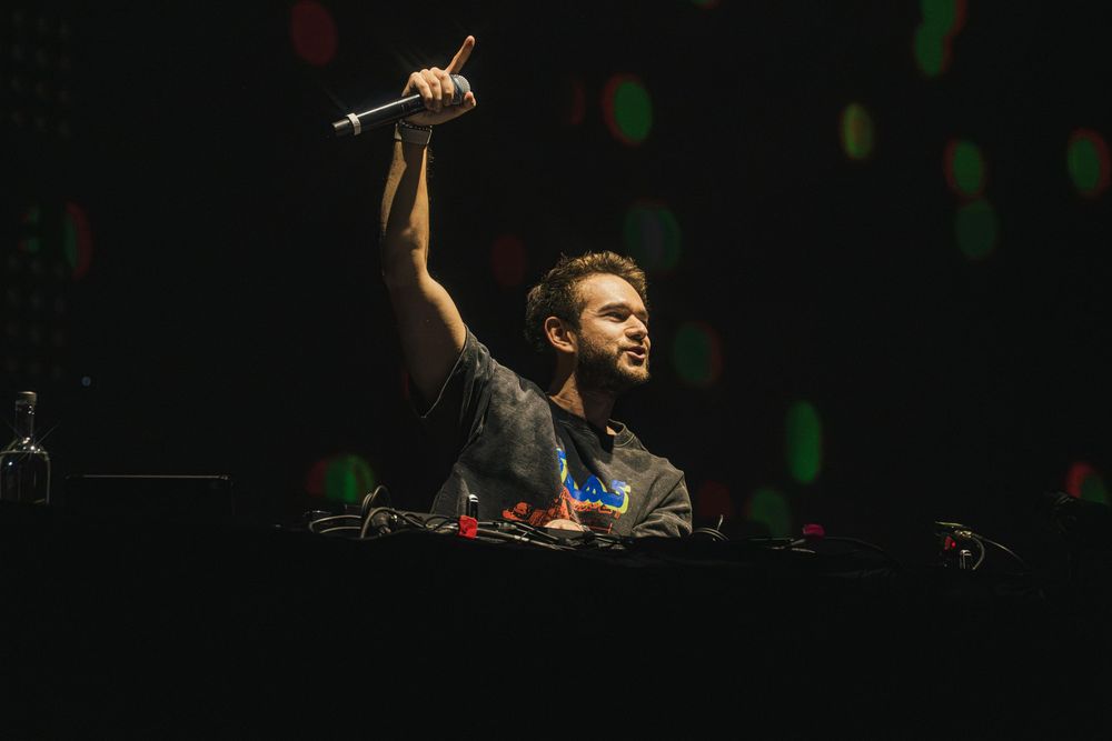 DJ performs behind decks at a concert with raised arm holding microphone against dark background with colored lights.