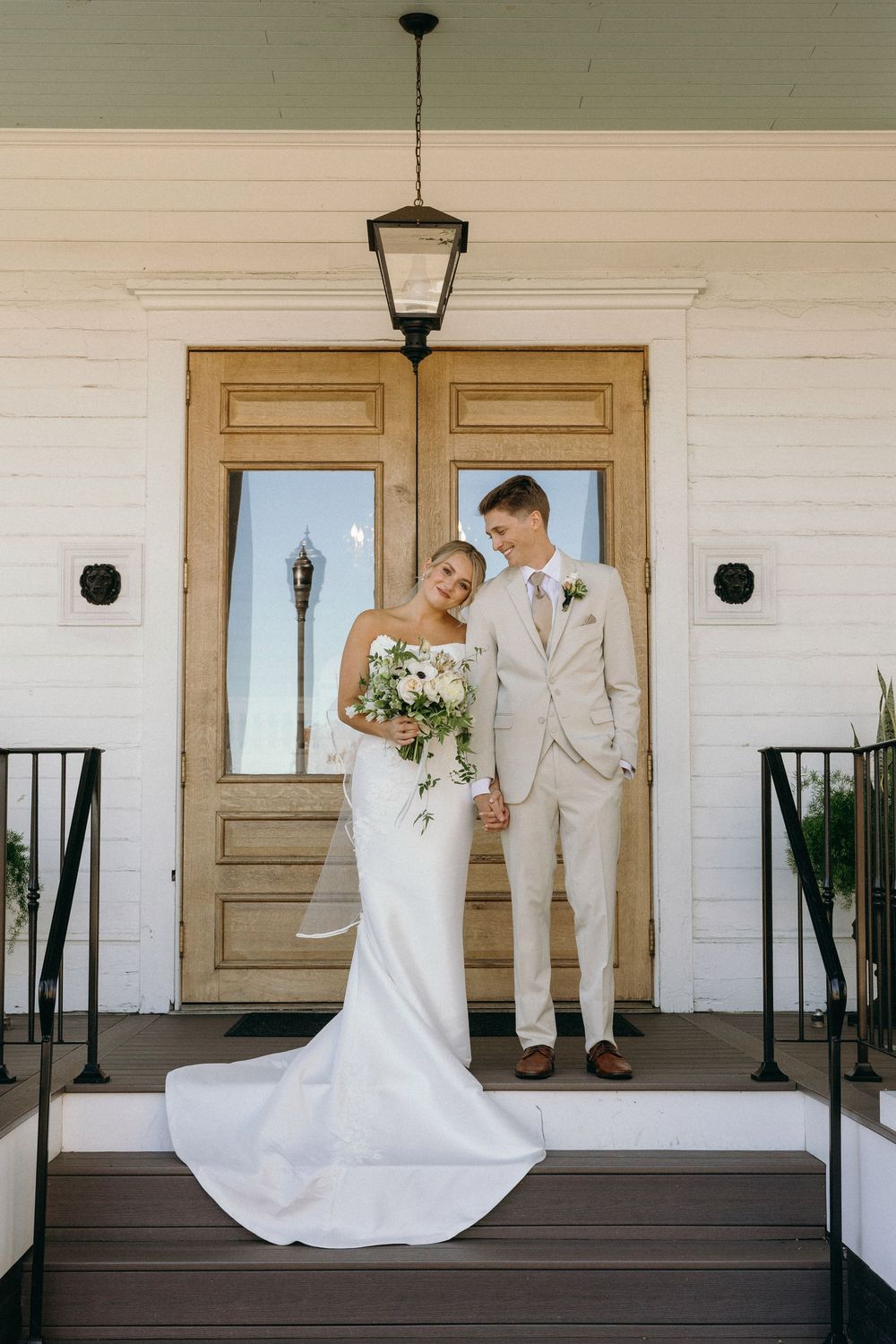A wedding couple poses together on a white porch with wooden doors in elegant white and beige attire at Lillian Gardens in Newnan, Georgia