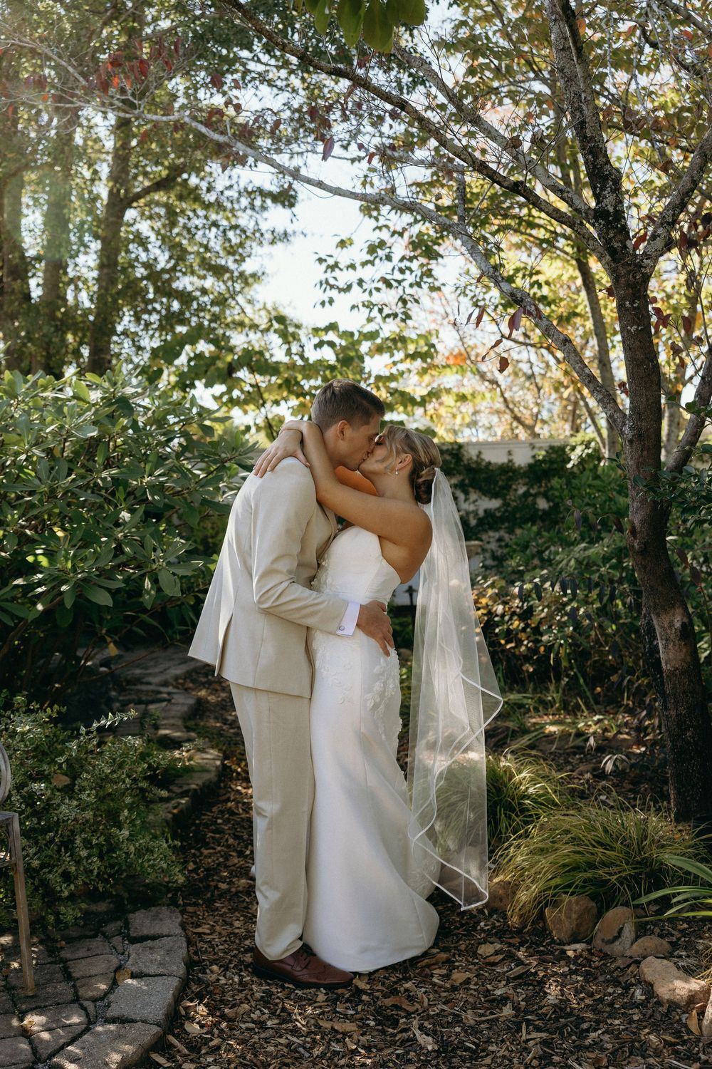 A romantic wedding moment captured in a garden setting with sunlight filtering through trees at Lillian Gardens in Newnan, Georgia